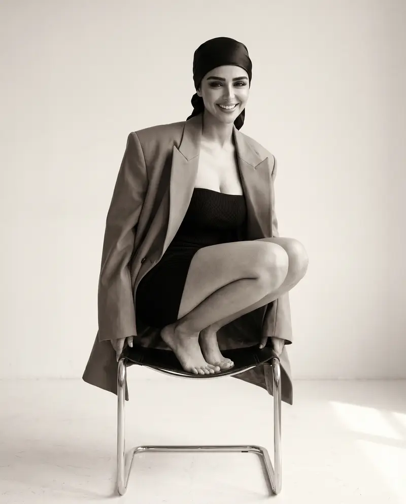 Crouched barefoot on a cantilever chair in an oversized blazer and headscarf